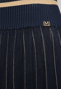 Námornícky plisovaný sukňa se zlatým metalickým vláknem, disponující žebrovaným pasem a zlatým logem jako akcentem. Texturovaná tkanina s vertikálními pruhy.