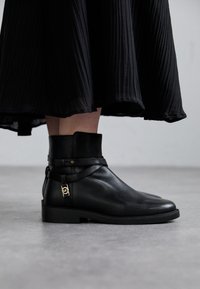 Černé kožené kotníkové boty s hladkým povrchem, zdobené páskem a zlatými detaily. Nízký blokový podpatek a kulatá špička.