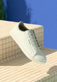 Baskets en cuir blanc avec une finition texturée, bout rond et coutures discrètes. Elles possèdent des lacets gris et une semelle en caoutchouc plate.