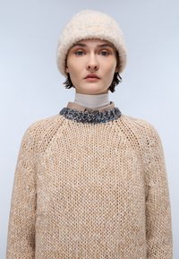 Beżowy sweter z dzianiny o teksturowanym, luźnym kroju i rękawami raglanowymi, z kolorowym żeberkowanym kołnierzem. Na górze miękka, kremowa, puszysta czapka.