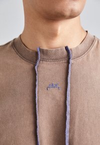 T-shirt en coton marron avec un col montant, des coutures bleues et un détail logo. Texture lisse avec une coupe décontractée et un design minimaliste.