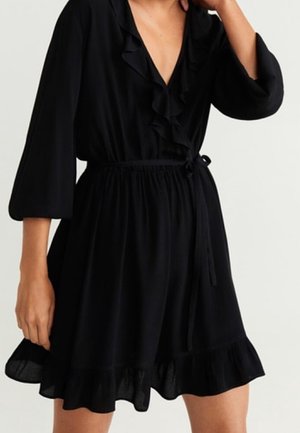 Schwarzes knielanges Kleid mit 3/4-Ärmeln, gerafftem V-Ausschnitt, Taillengürtel und gerafftem Saum, getragen von einer Person mit der Hand nahe am Hals.