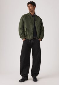 Χακί πράσινο μπουφάν μπόμερ με ριμπ μανσέτες, συνδυασμένο με φαρδιά μαύρα τζιν και μαύρα παπούτσια. Λείο ύφασμα, άνετη εφαρμογή και κλείσιμο με φερμουάρ.