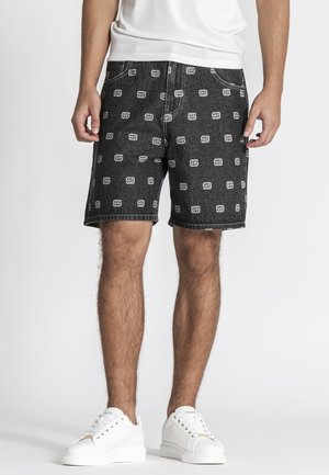 Mand iført sorte denimshorts med hvidt logomønster, hvide sneakers og en hvid t-shirt, stående mod en ensfarvet lys baggrund.