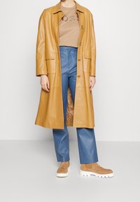 Długi, musztardowy skórzany płaszcz na beżowej koszulce z grafiką "BOSS", zestawiony z niebieskimi skórzanymi spodniami i brązowymi botkami na kostkę z białymi podeszwami.