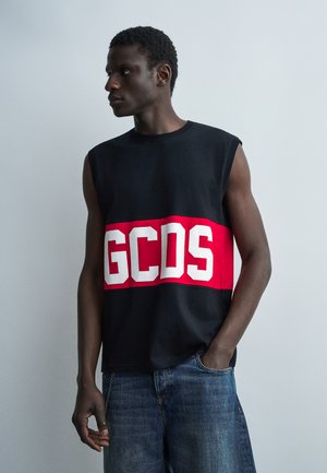 Άνδρας που φορά μαύρο μπλουζάκι χωρίς μανίκια με κόκκινη ρίγα και λευκά γράμματα "GCDS", σε συνδυασμό με μπλε τζιν σορτς, στέκεται σε απλό φόντο.