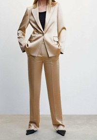 Béžový saténový oblek s upraveným sakem a kalhotami s rovnými nohavicemi. Tkanina má hladkou strukturu, obsahuje jeden knoflík a detaily kapes.