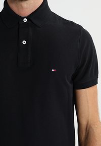 Musta polo-paita, joka on valmistettu tekstuurikankaasta, jossa on kolmen napin nappilista, lyhyet hihat ja pieni logo punaisilla, valkoisilla ja sinisillä korostuksilla.