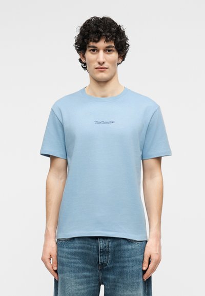 Νέος άνδρας με σγουρά μαλλιά που φοράει γαλάζιο t-shirt με το λογότυπο "The Kooples" και μπλε τζιν, στέκεται μπροστά σε απλό λευκό φόντο.