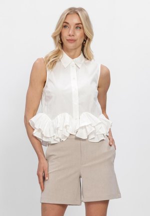 Blonde woman wearing sleeveless white blouse with ruffle hem and beige knee-length shorts, standing against plain background.