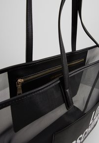 Svart tote-väska med genomskinlig meshkropp, läderdetaljer och en zippered inre ficka med gulddetaljer och varumärkeslogotyp.