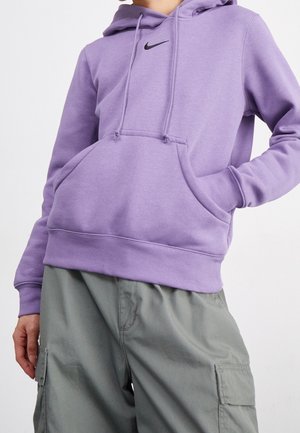 Osoba ubrana w jasnofioletową bluzę z kapturem Nike z przednią kieszenią oraz szare spodnie typu cargo na jednolitym białym tle.