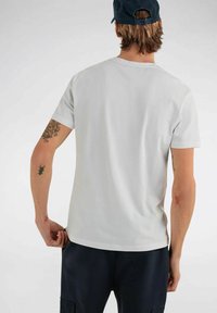 T-shirt grigio chiaro a maniche corte con scollo a giro, tessuto morbido e orlo leggermente curvo. Presenta tatuaggi visibili sulle braccia.