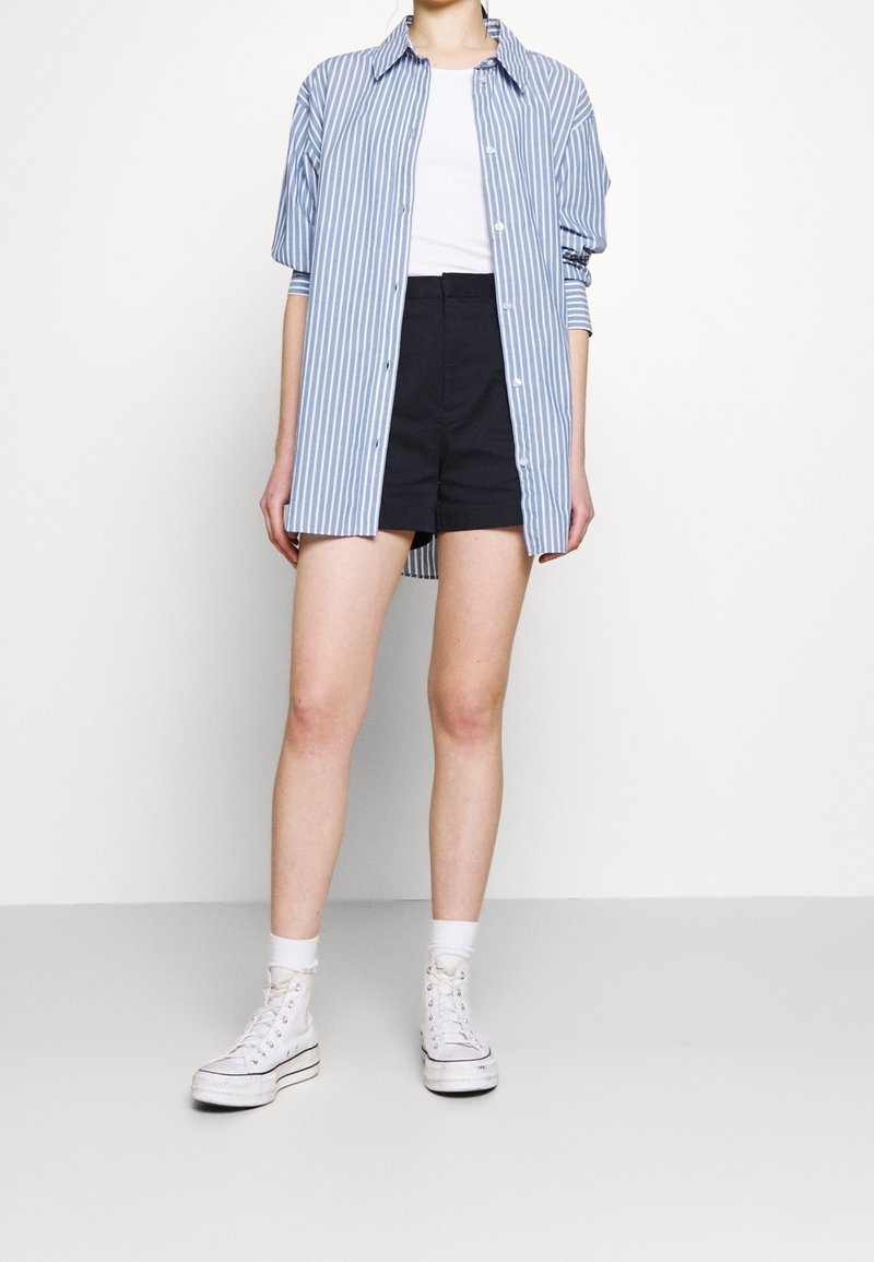 Camisa azul y blanca a rayas con botones, combinada con pantalones cortos negros. El modelo lleva zapatillas altas blancas y calcetines cortos. Conjunto fresco y casual.