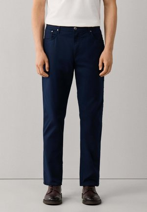 Man die donkerblauwe rechte broek, wit overhemd en donkerbruine leren schoenen draagt, staand tegen een effen achtergrond.