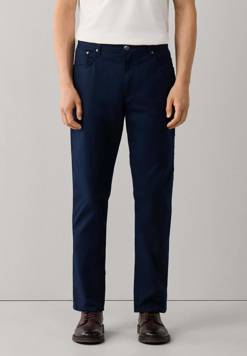 Hombre con pantalones de pierna recta de color azul oscuro, camisa blanca y zapatos de cuero marrón oscuro, de pie frente a un fondo liso.