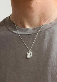 Collana con pendente d'argento caratterizzata da un charm rettangolare testurizzato, posizionata su una sottile catena, indossata sopra una maglietta grigia, che mette in evidenza un design minimalista.