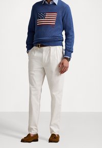 Pull en maille bleu avec motif de drapeau rouge et blanc, pantalon blanc, mocassins marron et une montre. La tenue présente des textures et des couleurs contrastées.