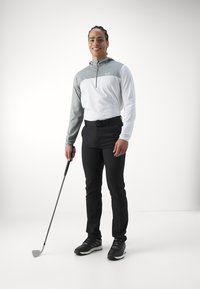 Tenue de golf comprenant un zip gris et blanc à manches longues, un pantalon noir et des chaussures de golf noires, avec un club de golf à la main.