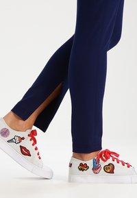 Sneakers bianchi con lacci rossi e patch ricamati; leggings blu navy con spacchetti laterali, tessuto liscio, design aderente e cuciture accentuate.