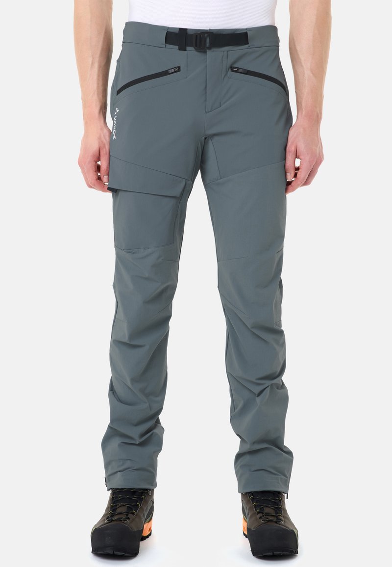 Pantalon d'extérieur gris avec ceinture noire, poches zippées et à rabat, porté avec des chaussures de randonnée noires et une chemise blanche.