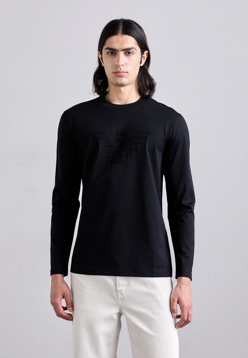 Garā piedurkņu melna T-kreklu, kas izgatavots no kokvilnas, ar diskrētu toņu logotipa dizainu uz priekšpuses. Vienkārša apaļa kakla izgriezums un pielāgotas piedurknes.