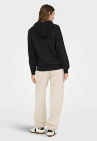 Czarny sweatshirt z kapturem, ze ribbowymi mankietami i dołem, w zestawieniu z beżowymi spodniami dresowymi i czarno-białymi sneakersami. Gładka tekstura materiału.