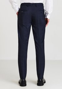 Mann in schmal geschnittenen, navyblauen karierter Hose und schwarzen Dressschuhen, stehend vor einem hellen, einfarbigen Hintergrund.