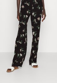 Black floral pants feature a pattern of pink flowers and green leaves. Paired with white slide sandals, highlighting a relaxed, casual style.