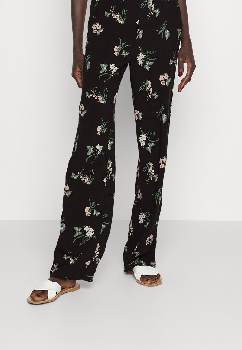 Black floral pants feature a pattern of pink flowers and green leaves. Paired with white slide sandals, highlighting a relaxed, casual style.
