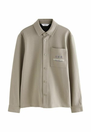 Beige button-up shirt made of soft fabric. Features a chest pocket with embroidered text and a classic collar design.