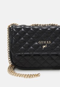 Bolso bandolera negro de piel sintética acolchada con una cadena dorada como correa y el logo dorado "GUESS girl" en el frente. Cuenta con cierre de solapa.