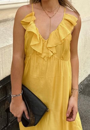 Vrouw draagt een gele, gerimpelde mouwloze jurk en houdt een zwarte designerk clutch met gouden kettingketting en armbanden vast.