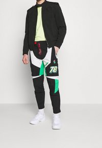 Černá bunda s vyvýšeným límcem, zelené tričko a kalhoty s barevnými bloky v černé barvě se bílými a zelenými akcenty. Model má na sobě bílé tenisky.