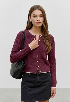Jeune femme aux longs cheveux bruns porte un cardigan bordeaux en maille torsadée boutonné, une jupe noire, des boucles d'oreilles en perles et porte un sac à bandoulière noir.