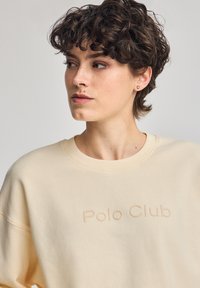 Maglioncino color crema con scollo rotondo e spalle cadenti, caratterizzato da un logo "Polo Club" in rilievo sul davanti. Tessuto morbido al tatto.