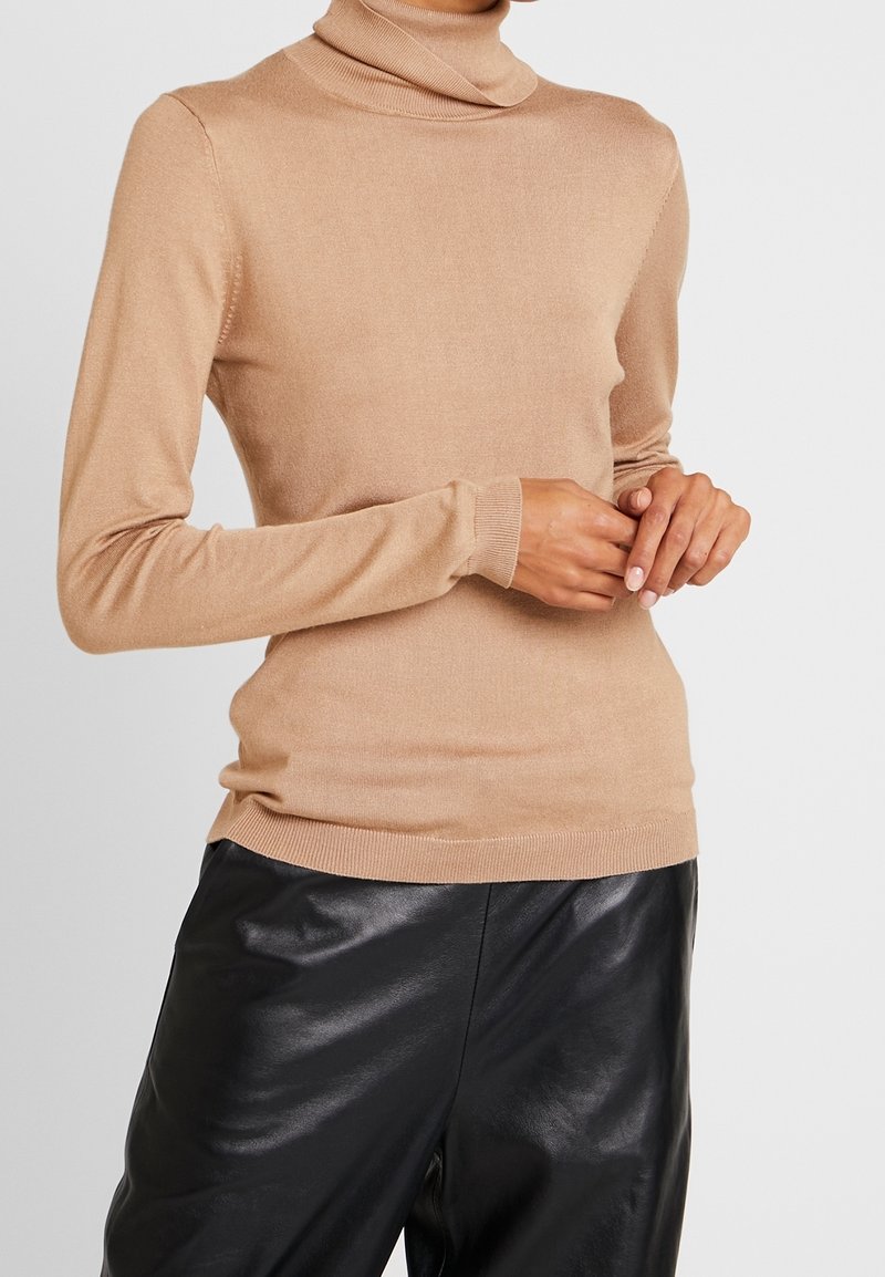 Personne portant un pull beige à col roulé à manches longues et un pantalon en cuir noir, les mains jointes devant sur un fond uni.
