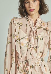 Blusa floral de tela suave en rosa claro, con volantes en el escote y las mangas, que presenta un lazo en el cuello y una cintura ceñida con cinturón.