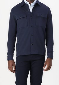 Marinblå skjortjacka i mjukt tyg med knappstängning framtill, två bröstfickor med lock och knappknäppta ärmslut. Ren, minimalistisk design.