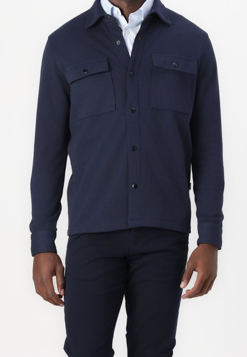 Marinblå skjortjacka i mjukt tyg med knappstängning framtill, två bröstfickor med lock och knappknäppta ärmslut. Ren, minimalistisk design.