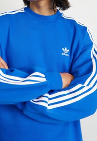 Sininen collegepaita, jossa korkea kaulus, pitkät hihat ja valkoiset kolmiraidat hartioilla. Adidas-logo rinnassa.