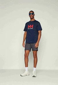 Άντρας που φοράει navy μπλούζα Helly Hansen, σκούρες βερμούδες, λευκές κάλτσες, λευκά αθλητικά παπούτσια και μεγάλα ανακλαστικά γυαλιά ηλίου, στέκεται σε απλό φόντο.