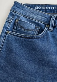 Jeans de denim en azul medio con un ligero desgaste, que cuentan con un bolsillo frontal, herrajes metálicos y una etiqueta que dice "MOTION FLEX" en la cintura.