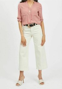Donna che indossa una blusa rosa polveroso, pantaloni bianchi a gamba larga a vita alta, una cintura nera con fibbia dorata e scarpe con tacco aperte color crema.