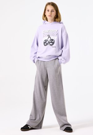 Lila Kapuzenpullover mit grafischen Kirschen, kombiniert mit grauen weiten Hosen mit einem subtilen Streifenmuster. Schwarze Slippers.