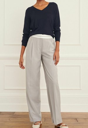 Kvinde iført en sort V-hals sweater, lysegrå plisserede bukser med vid ben, hvid undertrøje og cremefarvede ballet flats med sløjfe.