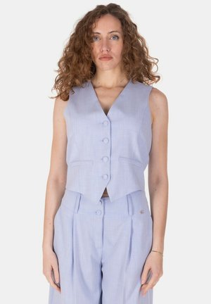 Donna con capelli ricci che indossa un gilet senza maniche azzurro chiaro abbottonato e pantaloni a vita alta coordinati, in piedi davanti a uno sfondo semplice.