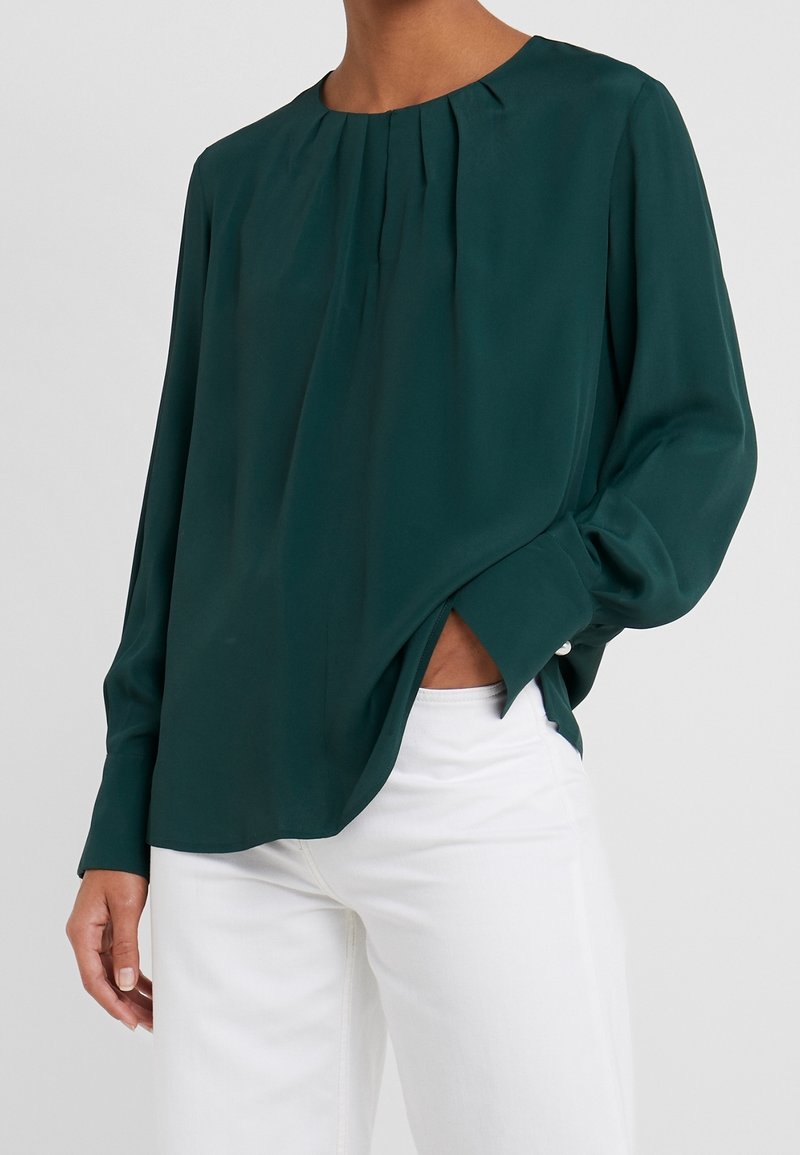 Blusa a maniche lunghe di colore verde scuro con scollo rotondo, dettaglio plissettato nella parte frontale e vestibilità rilassata, abbinata a pantaloni bianchi.