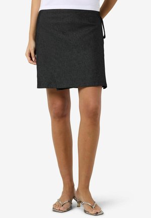 Kvinne iført et svart omslagskjørt og heeled sandaler med slangeskinnmønster, stående foran en hvit bakgrunn.