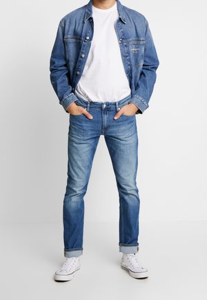 Chaqueta vaquera de lavado claro, combinada con una camiseta blanca y jeans azules. Los jeans son ajustados con puños doblados; los zapatos son zapatillas deportivas blancas.